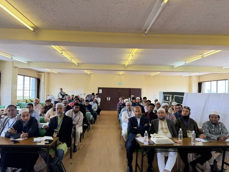 Rows of men, many wearing kufi caps and lanyards, seated at tables in a community hall, listening attentively as a meeting takes place.