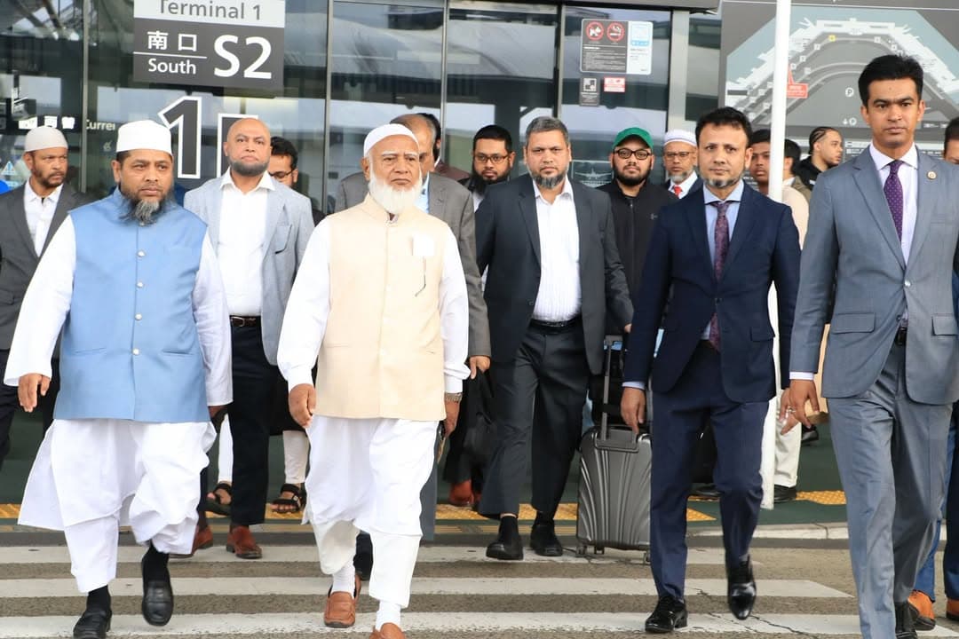 A group of men in traditional and formal attire walk out through the South Exit S2 doors of Terminal 1 at an airport, with an elderly man in a white cap and beige vest walking beside another man in a blue vest at the front, followed by several others in suits, one pulling a rolling suitcase.