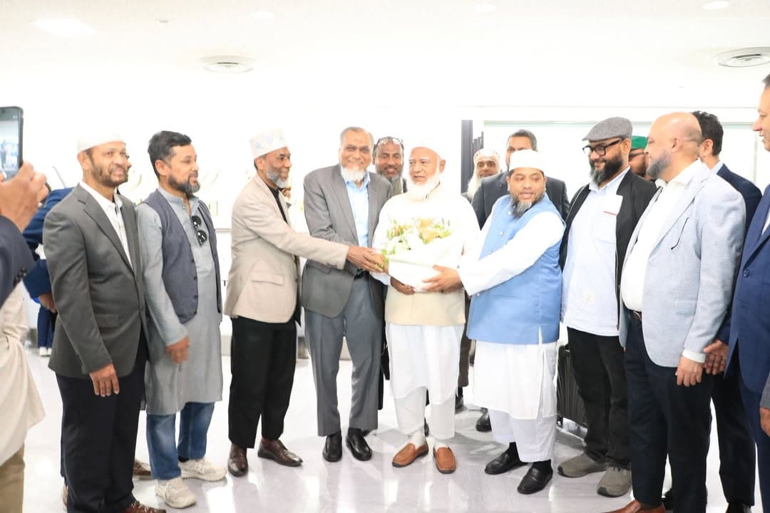 A group of men in an airport lobby gather around an elderly man in a white robe and cap, presenting him with a bouquet of flowers while others look on and one person films with a phone.