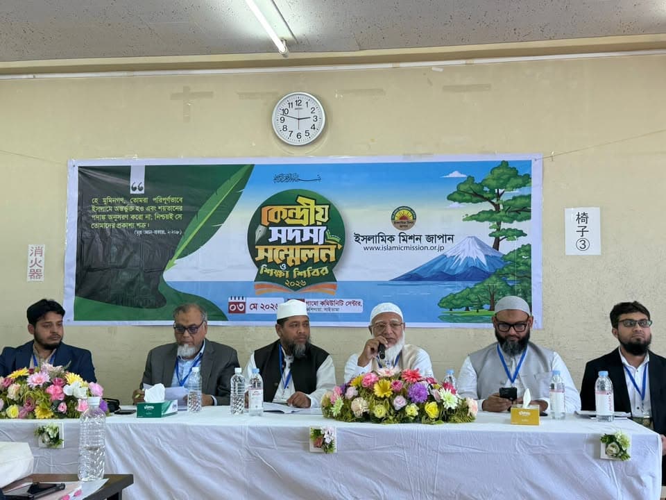 Six men sit at a table with floral arrangements and water bottles, one speaking into a microphone, in front of a banner reading "Central Members' Conference and Education Camp 2025" with Islamic Mission Japan's logo, in a room with a wall clock behind them.