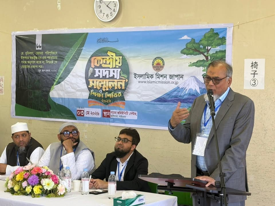 A man in a gray suit speaks at a podium microphone during a conference, with three seated men beside him at a flower-decorated table and a banner reading "Central Members' Conference & Education Camp" with Islamic Mission Japan's logo behind them.