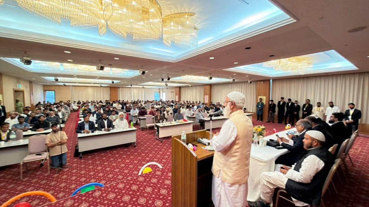 A speaker in traditional white attire and a cap addresses a large seated audience from a podium in a banquet hall, with several officials seated at a head table beside him and staff standing along the back wall.