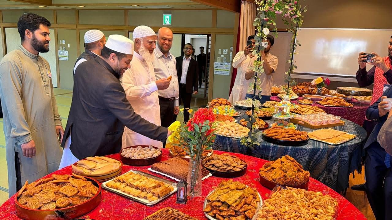 A group of men in traditional attire gather around a table laden with an array of sweets, pastries, and snacks decorated with flowers, while people in the background take photographs.