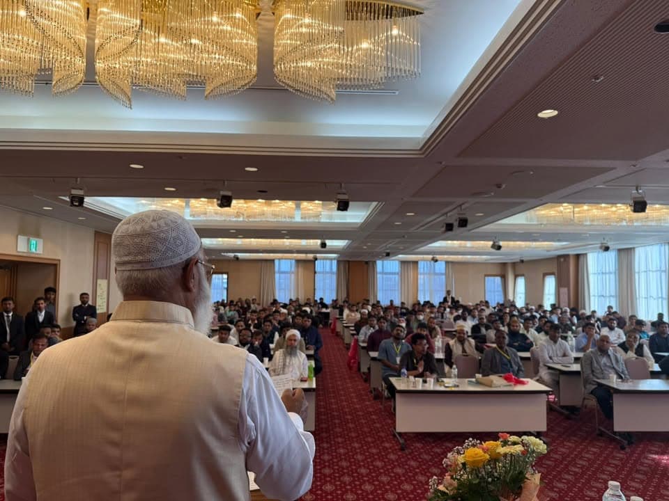 A man wearing a white cap and vest stands at the front of a large banquet hall, speaking and holding papers, while a large seated audience of men fills rows of tables under a decorative chandelier.