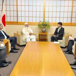 A man in traditional white robes and a turban sits in a meeting room in Japan, engaged in conversation with Japanese officials in suits seated around a low table, with a Japanese flag and a flower arrangement in the background.