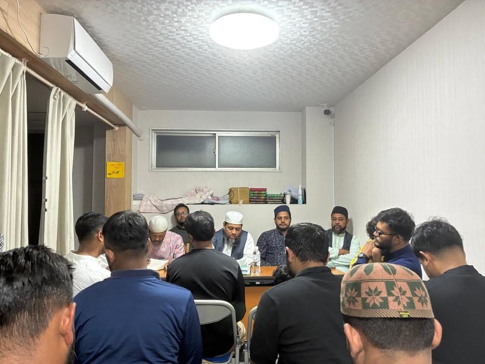 A group of men, some wearing prayer caps, sit around a table in a small room with a curtained window and shelf of books, gathered for a meeting.