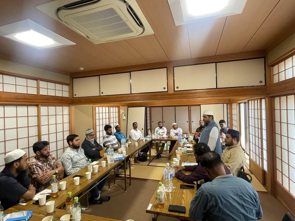 A group of men, some wearing caps and traditional attire, sit around low tables arranged in a U-shape in a tatami-floored Japanese-style room, listening to a man standing and speaking at the front.