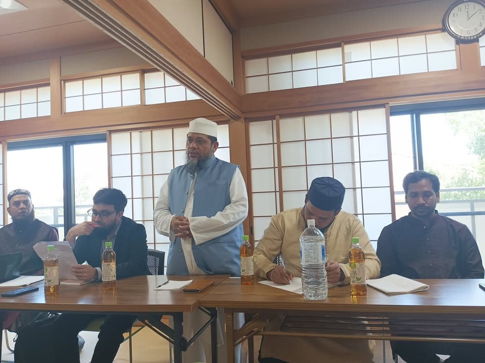A man in a white cap and light blue vest stands and speaks at a wooden table in a tatami-floored room with sliding paper doors, while several other men sit beside him, some writing on papers, with tea bottles placed on the table.