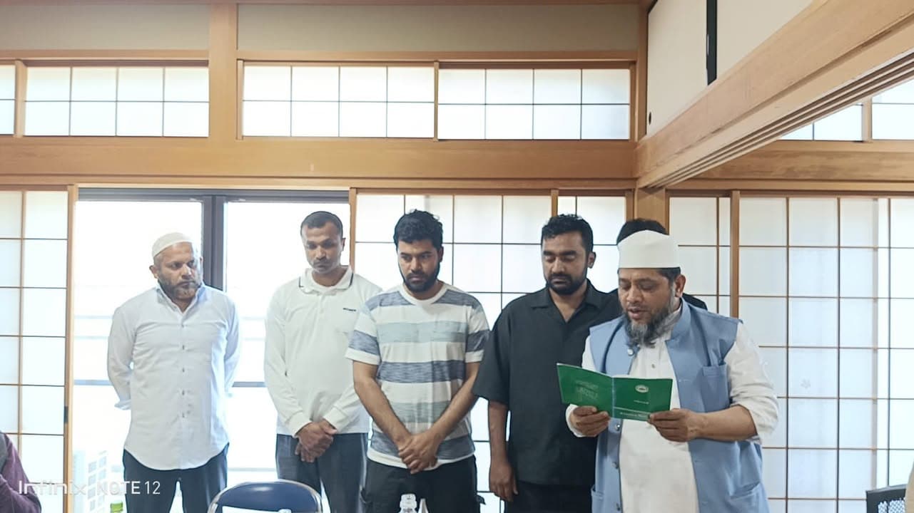 Five men stand in a row inside a tatami-style room with paper sliding doors, hands clasped in front of them, as the man on the right, wearing a white cap and blue vest, reads aloud from a small green booklet.