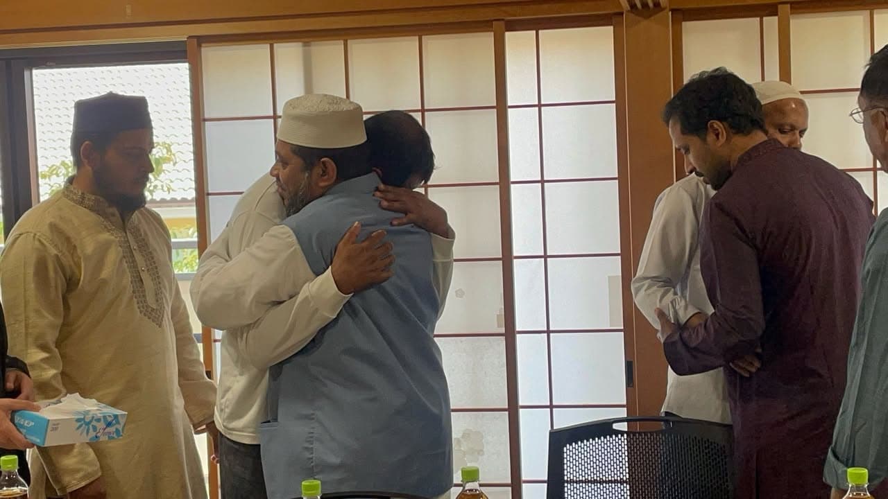 Two men embrace warmly in a traditional Japanese-style room with shoji screens, while several other men in kurta and thobe stand around them, some holding water bottles and a tissue box.