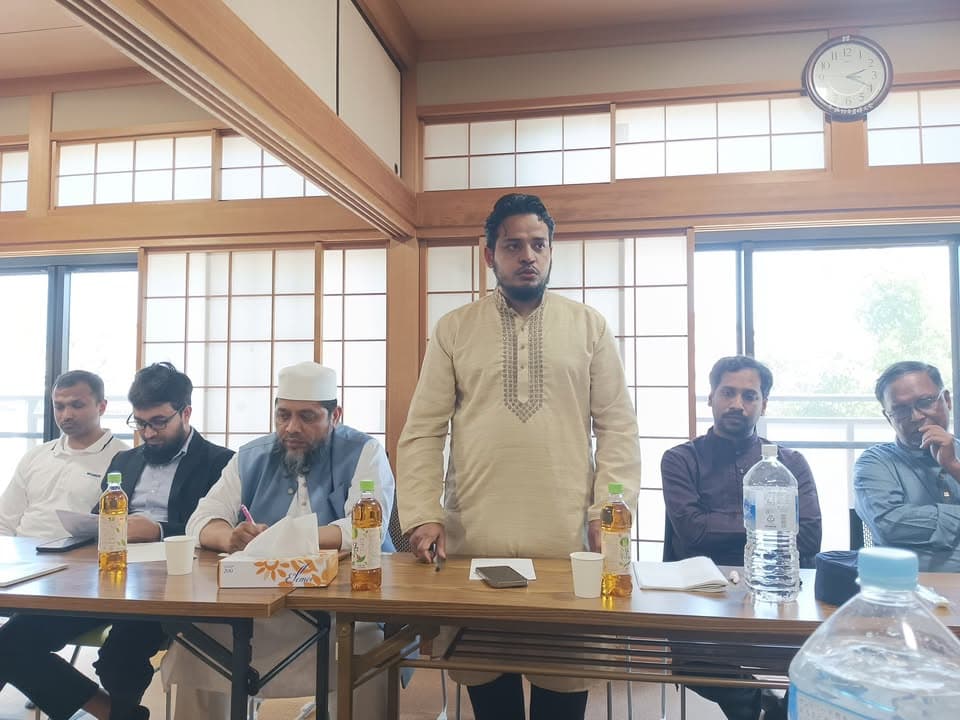 A man in a light beige kurta stands and speaks while seated men, including one wearing a white cap, listen around a table in a tatami-style room with sliding paper doors.