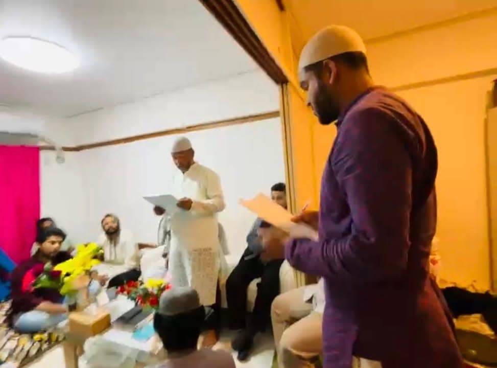 Men in traditional Islamic dress, some wearing prayer caps, gather in a room decorated with flowers, with two men standing and reading from papers as others sit and listen.