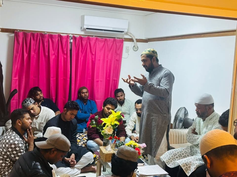 A man in a gray kurta and embroidered cap stands and speaks with his hands raised while a group of men in prayer caps sit on the floor around a low table with flowers, in a room with a pink curtain and white walls.