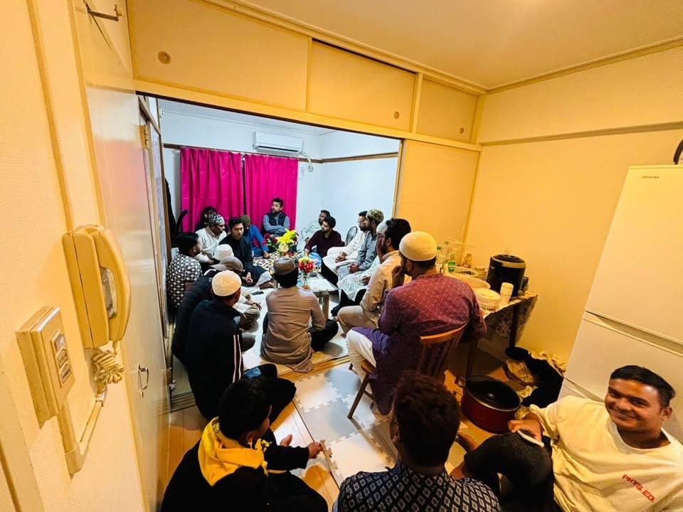 A group of men, many wearing traditional caps and kurtas, gather closely seated on the floor and chairs in a small apartment room to hold a meeting.