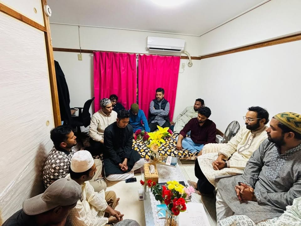A group of men in traditional Islamic attire sit together on the floor of a small room, gathered around a low table decorated with flower vases, engaged in discussion.