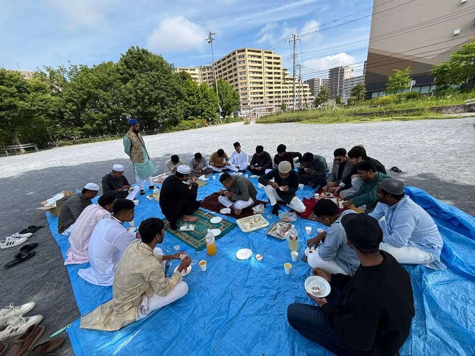 A large group of men in traditional Islamic clothing sit in a circle on blue tarps and prayer mats spread on a paved area in a park, eating from plates and cups, with apartment buildings and trees visible in the background.