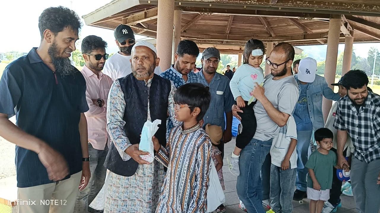 A group of Muslim men, children, and a woman holding a young girl gather under a wooden pavilion, with a boy in a striped kurta handing a small gift bag to an elderly man wearing a cap and vest.