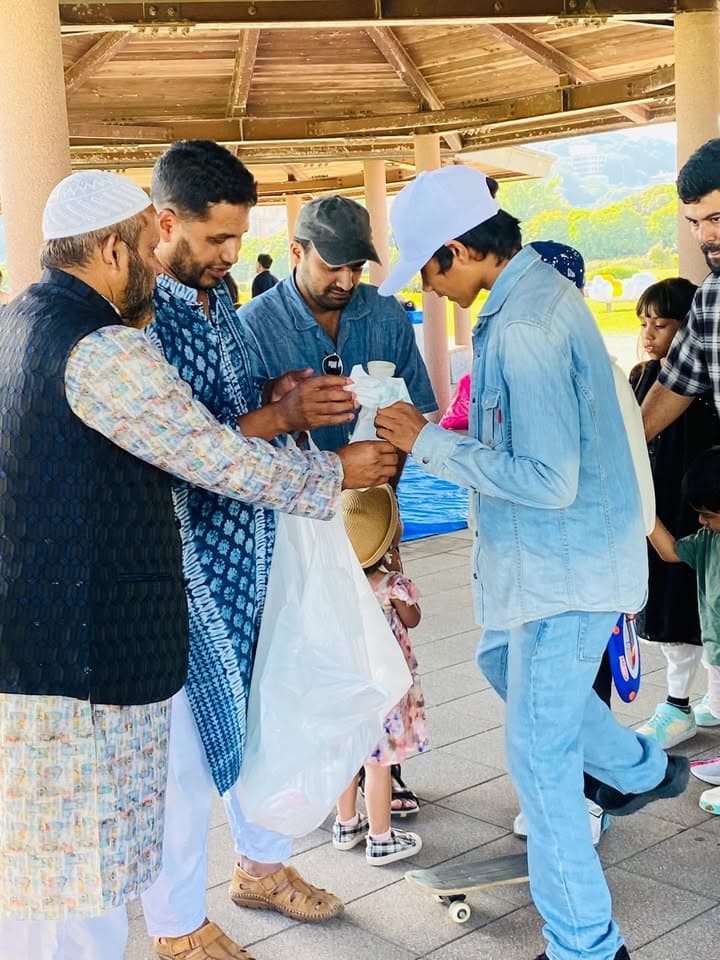 A group of men, some in traditional kurta and taqiyah caps, gather under a wooden pavilion sharing cups of drinks, with children and other families nearby.