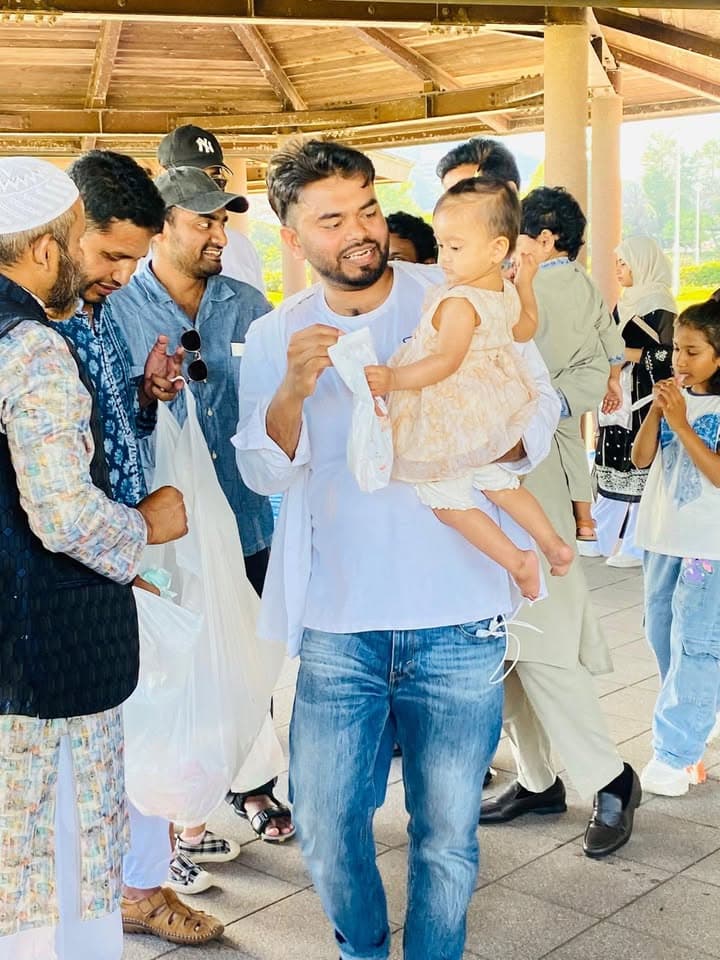 A man in a white shirt holds a young girl while standing under a wooden pavilion, surrounded by other men and families dressed in traditional clothing.