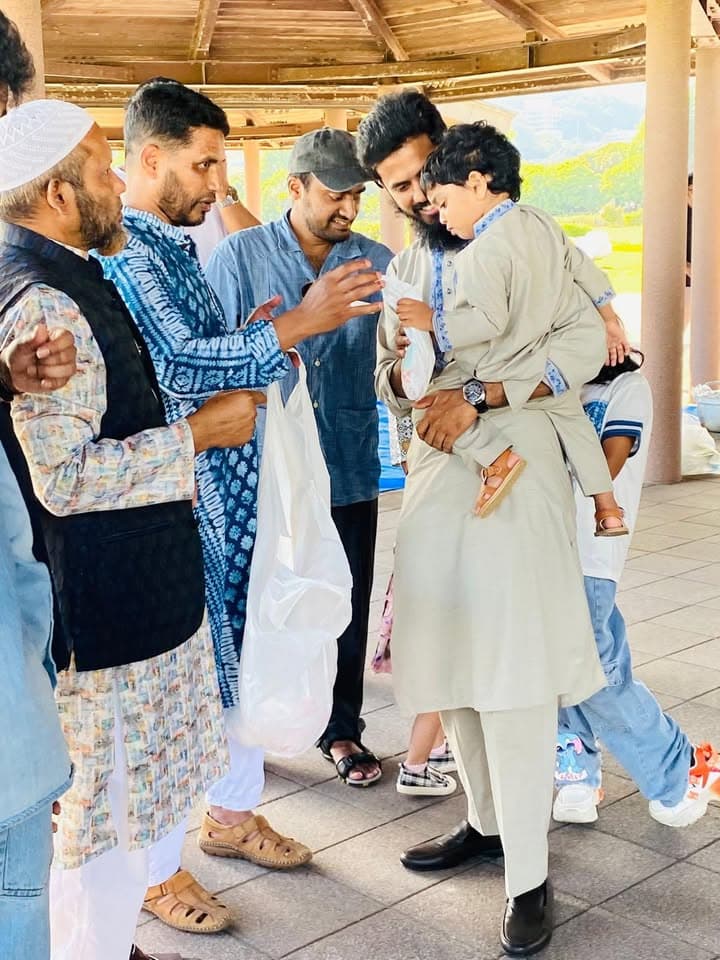 A group of men in traditional South Asian dress gather under a wooden pavilion outdoors, with one man holding a small child while others exchange items in plastic bags.