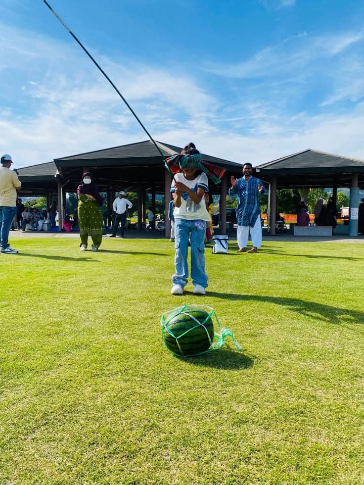 A blindfolded child holds a stick raised overhead, taking part in a watermelon-splitting game on a grassy lawn, with a netted watermelon placed on the ground in front, and families and a shelter pavilion in the background.