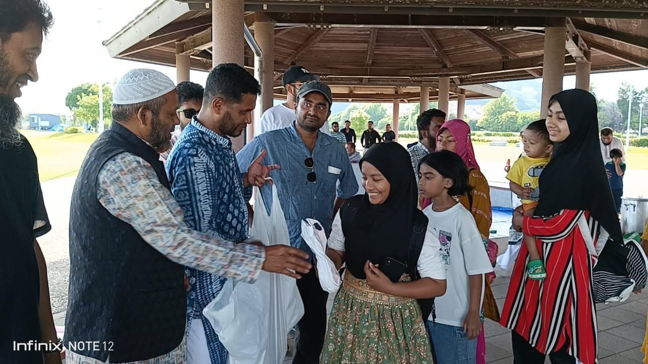 Under a wooden pavilion at a beachside park, men, women, and children gather as an elder in a white cap hands a bag to a young girl in a black hijab, with families in colorful traditional dress standing around them.