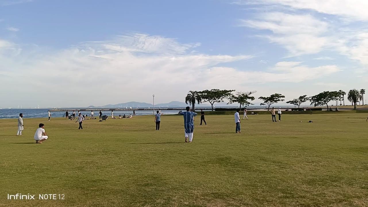 People in traditional South Asian clothing gather and stand on a wide grassy field beside the sea, with palm and other trees and distant hills in the background under a partly cloudy sky.