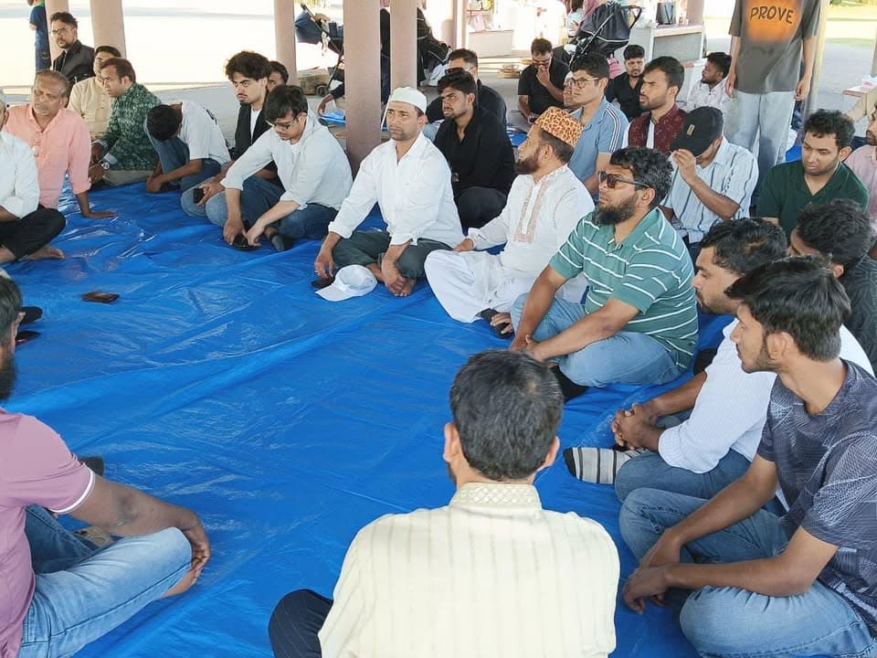 A large group of men sit cross-legged in a circle on a blue tarp under a shelter, listening attentively to someone speaking.