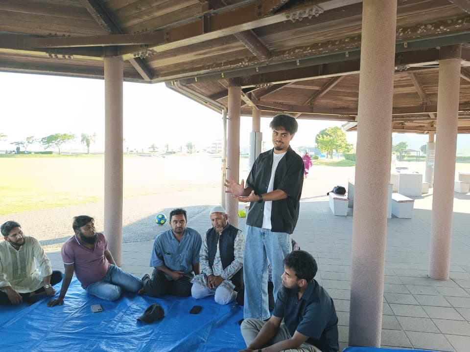 A young man stands and speaks while gesturing with his hands, addressing a group of men seated on a blue tarp under an open-sided pavilion near a grassy beach area.