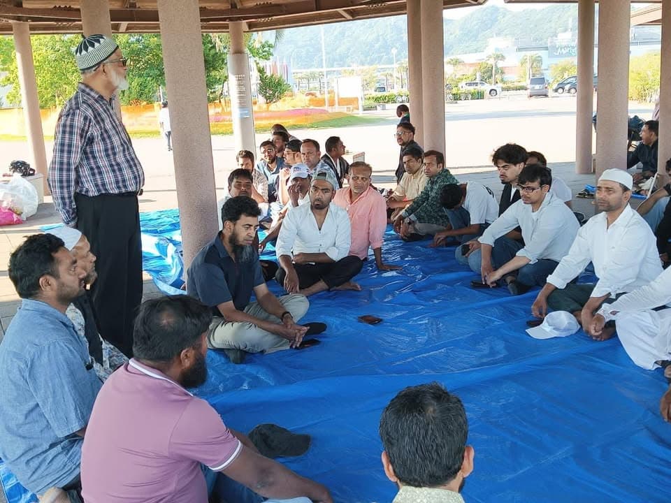 A group of men sit cross-legged on blue tarps spread under an outdoor pavilion, listening to an elderly man in a plaid shirt and skullcap who stands and speaks to them, with a seaside landscape visible in the background.