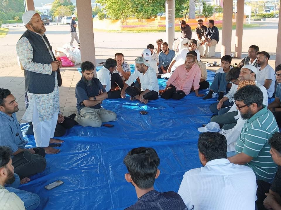 A group of men sit on blue tarps under an outdoor shelter while one man stands and speaks to them, with trees and parked cars visible in the background.