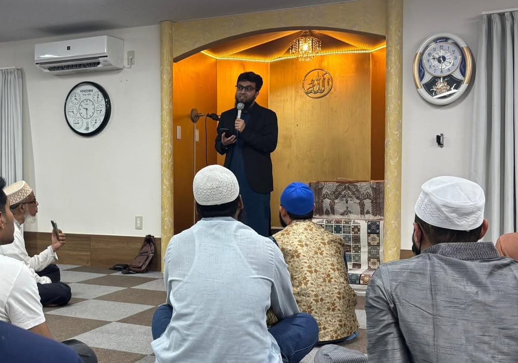 A man stands near the mihrab of a mosque, speaking into a microphone while a seated audience of men, some wearing prayer caps, listens attentively on the carpeted floor.