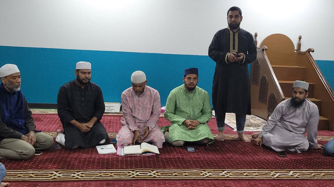 A group of men in traditional Islamic dress sit on a prayer carpet inside a mosque, with one man standing near a wooden minbar and another reading from a book placed on the floor.