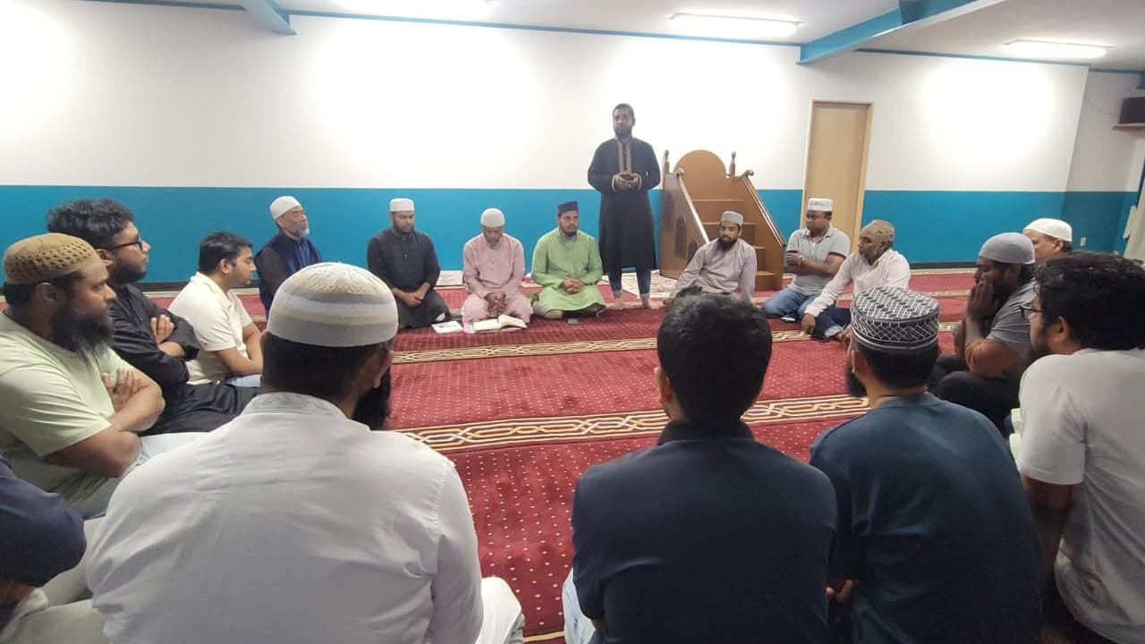 A man stands and speaks near the mosque's mimbar while a group of men sit on the red patterned carpet around him, listening attentively inside the prayer hall.