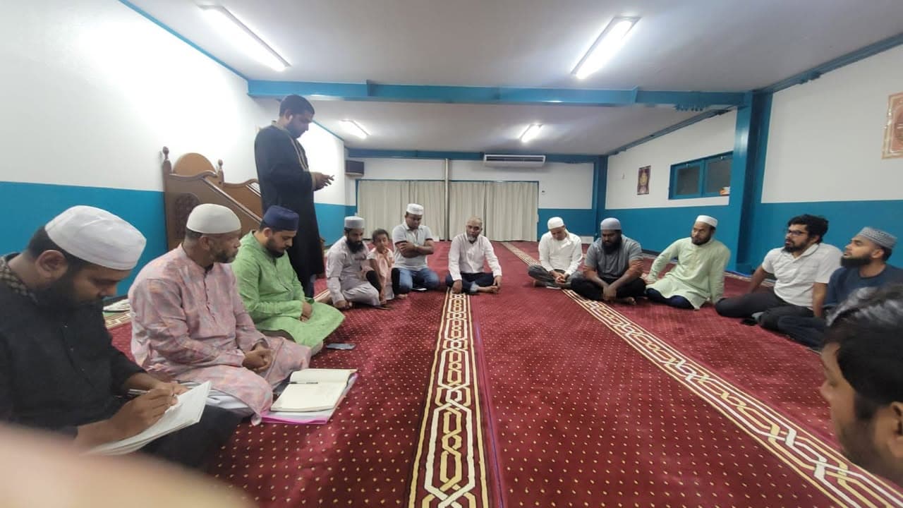 A group of men and one child sit cross-legged in two rows along a red patterned carpet inside a mosque prayer hall, some wearing white or colored kurtas and prayer caps, while one man stands speaking and two others sit taking notes on notebooks.