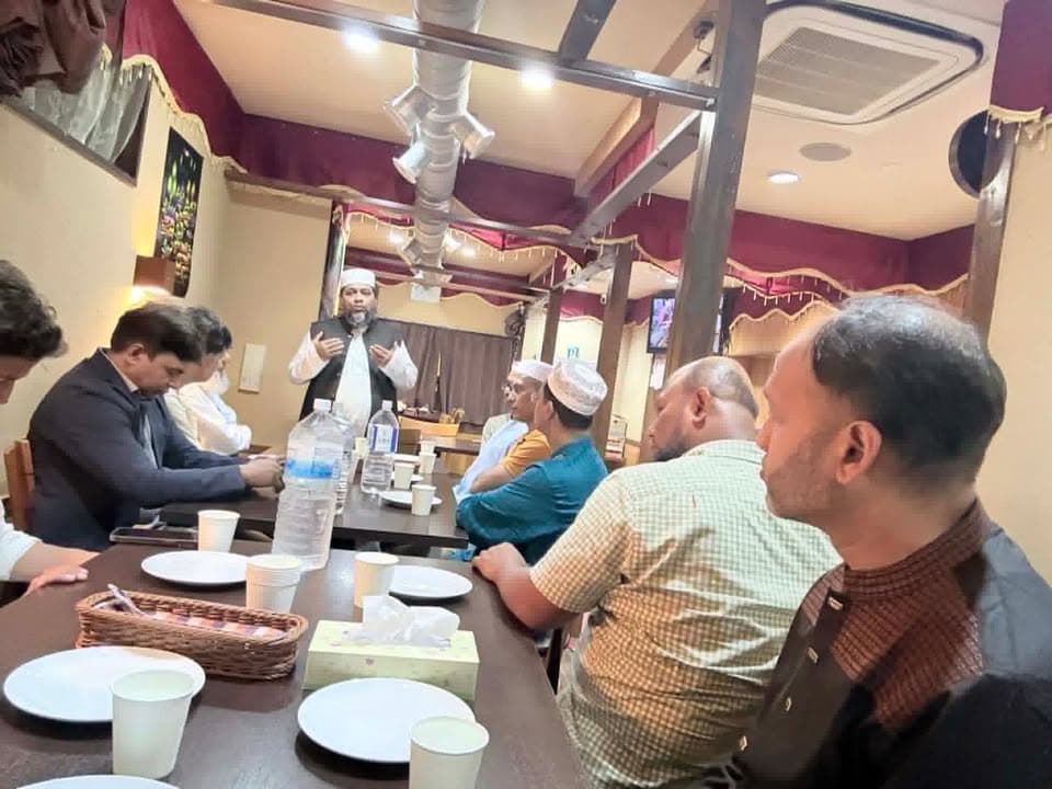 A man in a white cap and robe stands at the head of a table making a supplication with open palms, while a group of seated men listen with heads bowed around a table set with plates, cups, and water bottles in a restaurant with red drapery.
