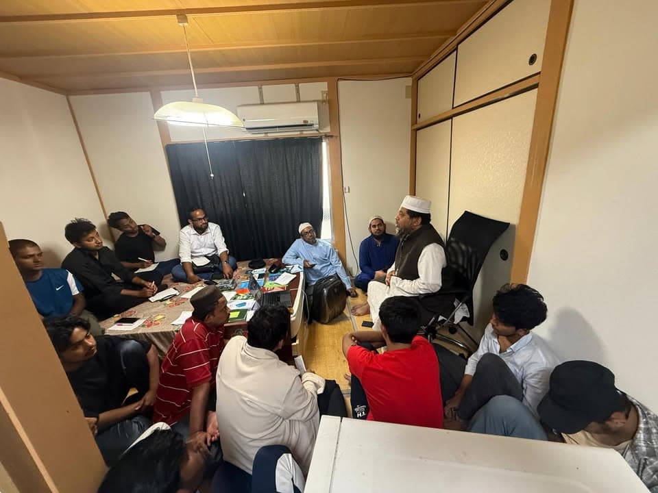 A group of men and young students sit closely together on a tatami floor in a small Japanese-style room, listening as two men in white caps speak to them.
