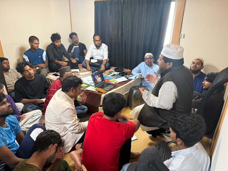 A group of men and young men sit closely together on a tatami-floored room, gathered around a low table with a laptop and papers, listening to a man in a turban and vest who is speaking and gesturing.