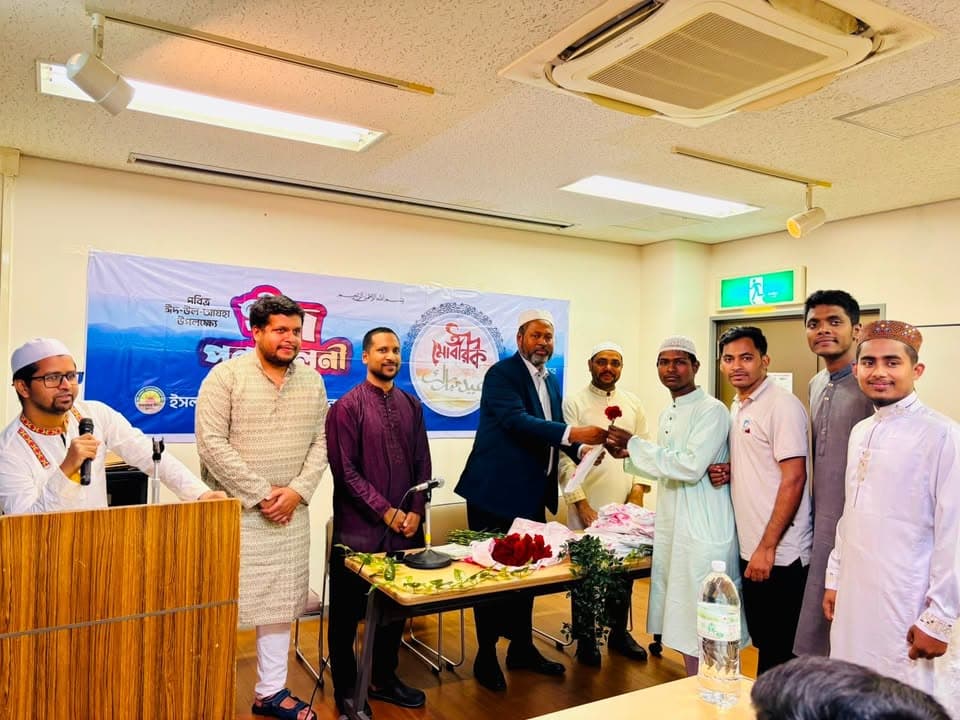 A group of men in traditional South Asian and Islamic attire stand together in a meeting room, with an older man in a suit handing a red rose and a certificate to a younger man in a white kurta, in front of a banner reading "Eid Mubarak" and a table with roses and flowers, while a man at a podium speaks into a microphone.