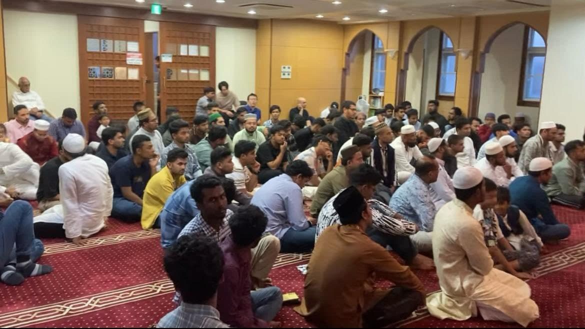 A large group of men and boys sit closely together on a red patterned carpet inside a mosque prayer hall, listening attentively to a speaker, with arched wooden doorways and windows in the background.