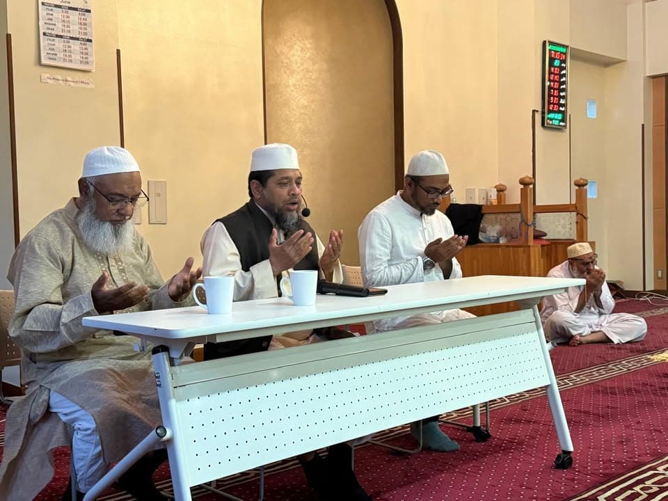 Four men in traditional white and cream robes with prayer caps sit around a white table inside a mosque prayer hall, hands raised in supplication, with prayer timetables and a digital clock displayed on the wall behind them.