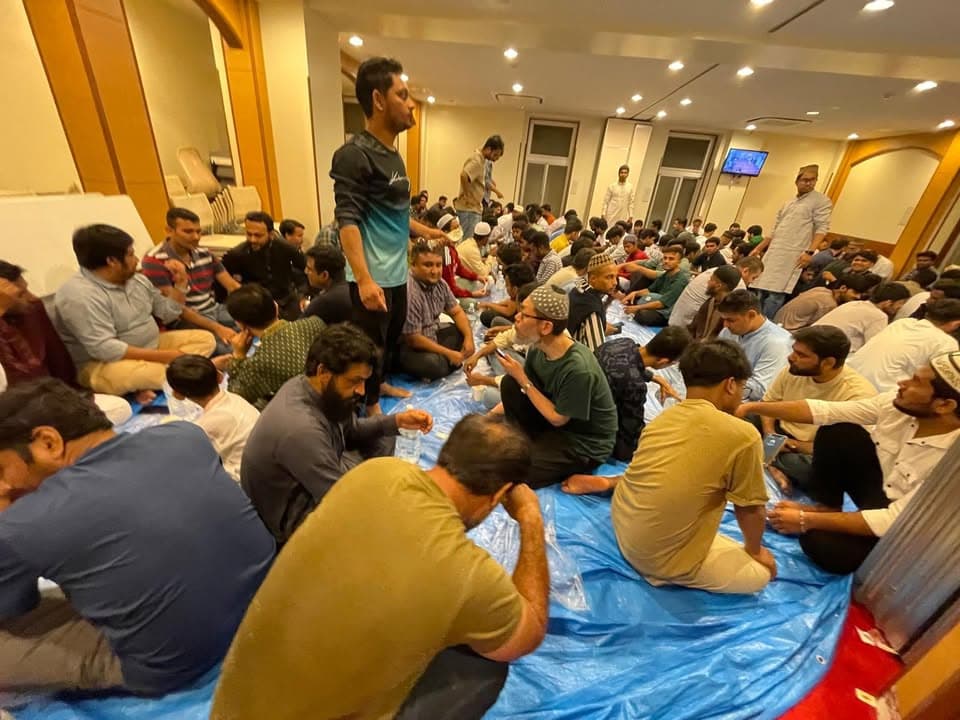 A large group of men sit closely together on blue tarp sheets spread across a mosque hall floor, listening and talking as bottled water is passed around, with more people gathered in the background under bright ceiling lights.
