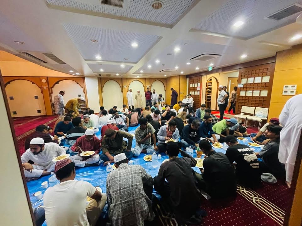 A large group of men sit on plastic sheets spread across the mosque floor, eating a meal together from plates while others stand and talk in the background of the prayer hall.