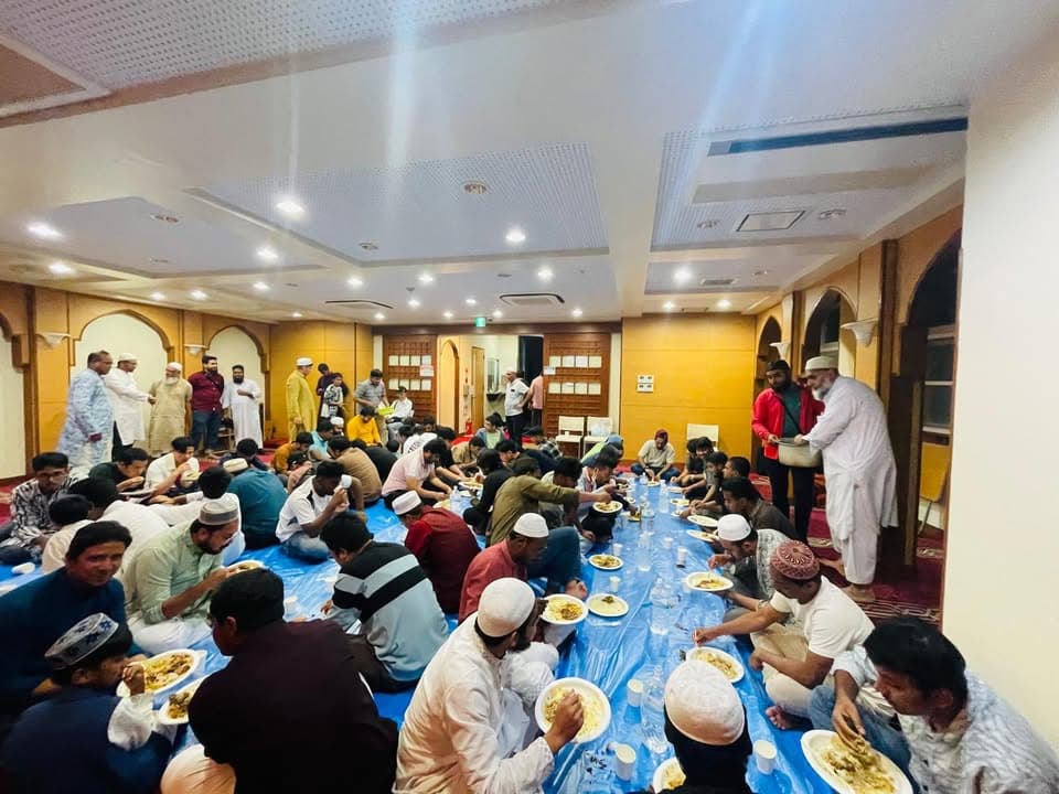 Rows of men sit on a blue tarp on the floor of a mosque hall, eating a meal together from plates while others stand and serve food nearby.