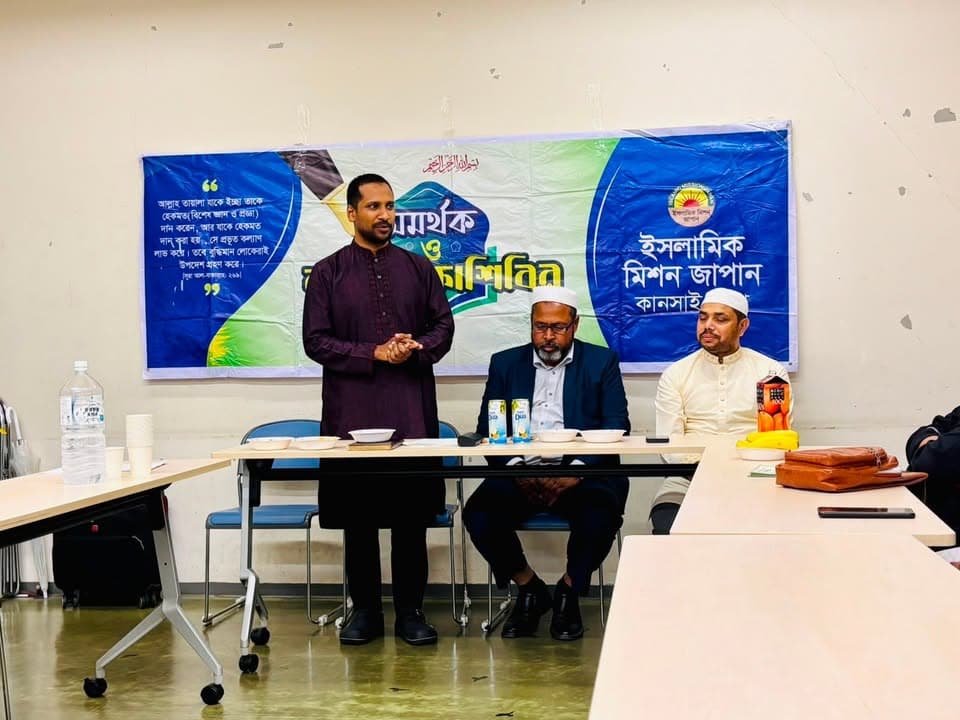 A man in a maroon kurta stands and speaks at a table while two seated men listen, in front of a large blue banner with Bengali text and the Islamic Mission Japan Kansai logo.