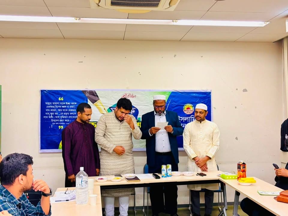 Four men stand in front of a Bengali-language banner in a meeting room, one reading from a paper while another wipes his eyes, with attendees seated at tables in the foreground.