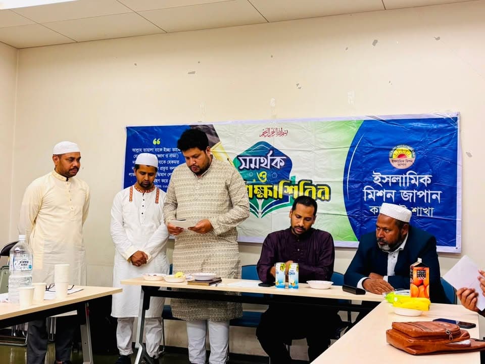 A man in traditional attire stands reading from a paper at a table, with two seated men and two standing men beside him, in front of a large banner in Bengali and Japanese reading "Islamic Mission Japan, Kansai Branch."