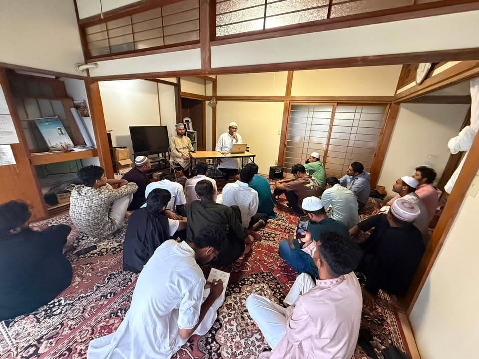 A group of men sit cross-legged on patterned carpets in a traditional Japanese-style room with tatami and sliding paper doors, listening to two speakers seated at a table at the front of the room, one of them using a laptop.