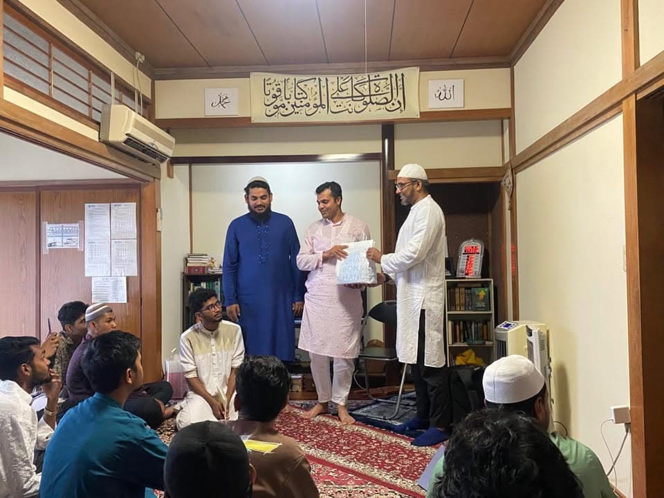 Inside a prayer hall with tatami-style flooring and Arabic calligraphy banners on the wall, three men in traditional Islamic dress stand while one hands a gift box to another, as a group of men and boys sit on the carpeted floor watching.