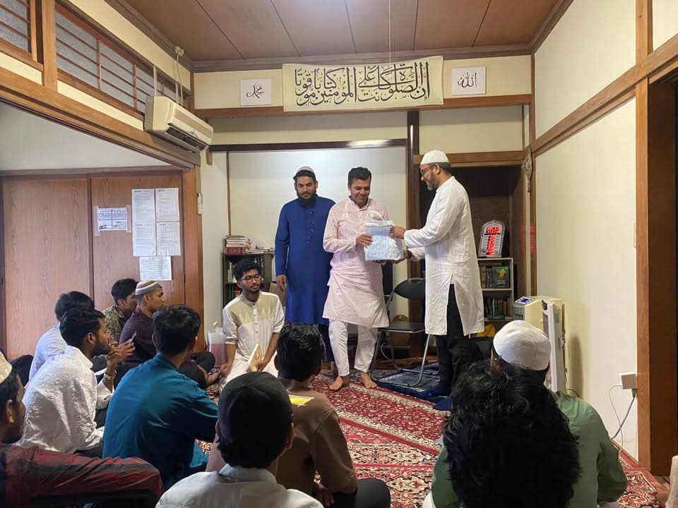 In a prayer room decorated with Arabic calligraphy, three men in traditional white and blue attire stand while one hands a wrapped item to another, as a group of men and boys sit on a patterned carpet listening.